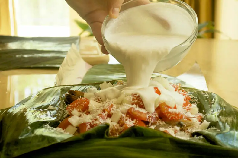 Pour coconut milk into a banana leaf stuffed with yam onion prawns and tomato 