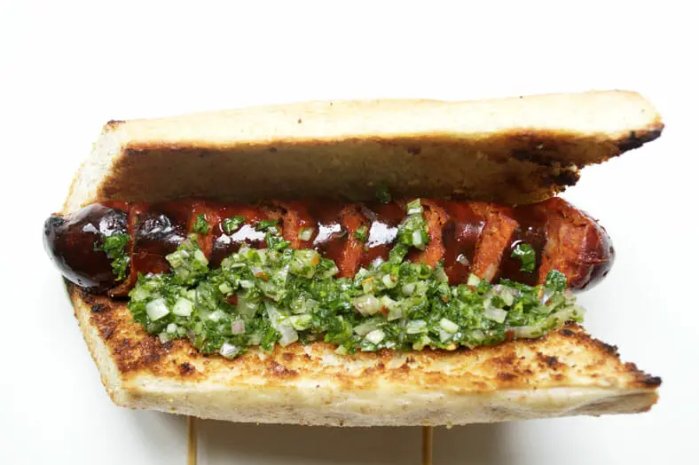 Grilled chorizo sausage inside a toasted baguette topped with a heaping dollop of chimichurri