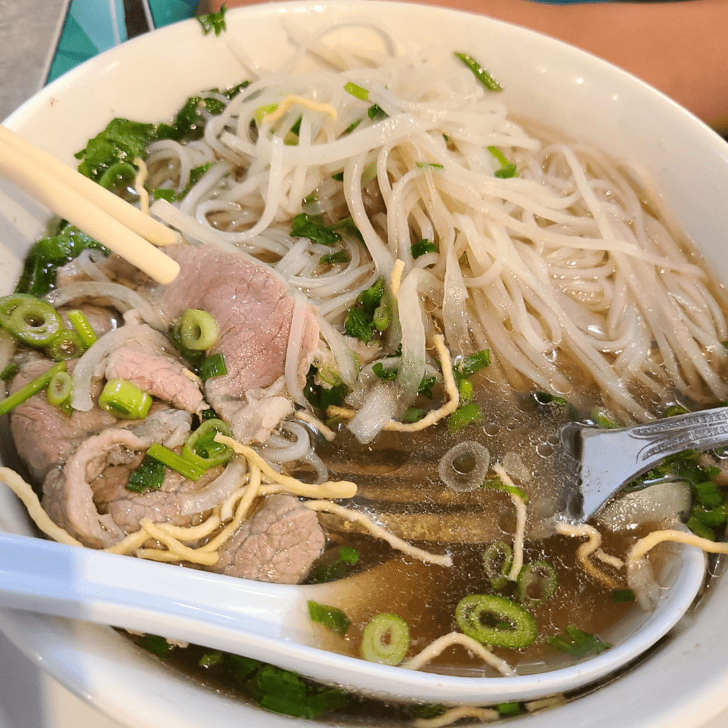 bowl of pho noodle soup with rare eye round steak chopsticks and a soup spoon
