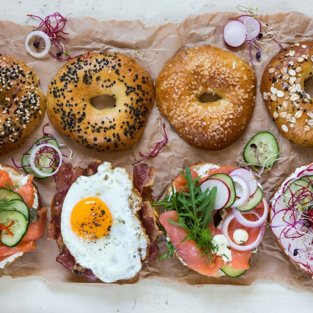 top view of open bagel sandwiches side by side. bagel sandwiches are filled with smoked salmon, cucumbers, onions, dill, eggs, bacon, and more.