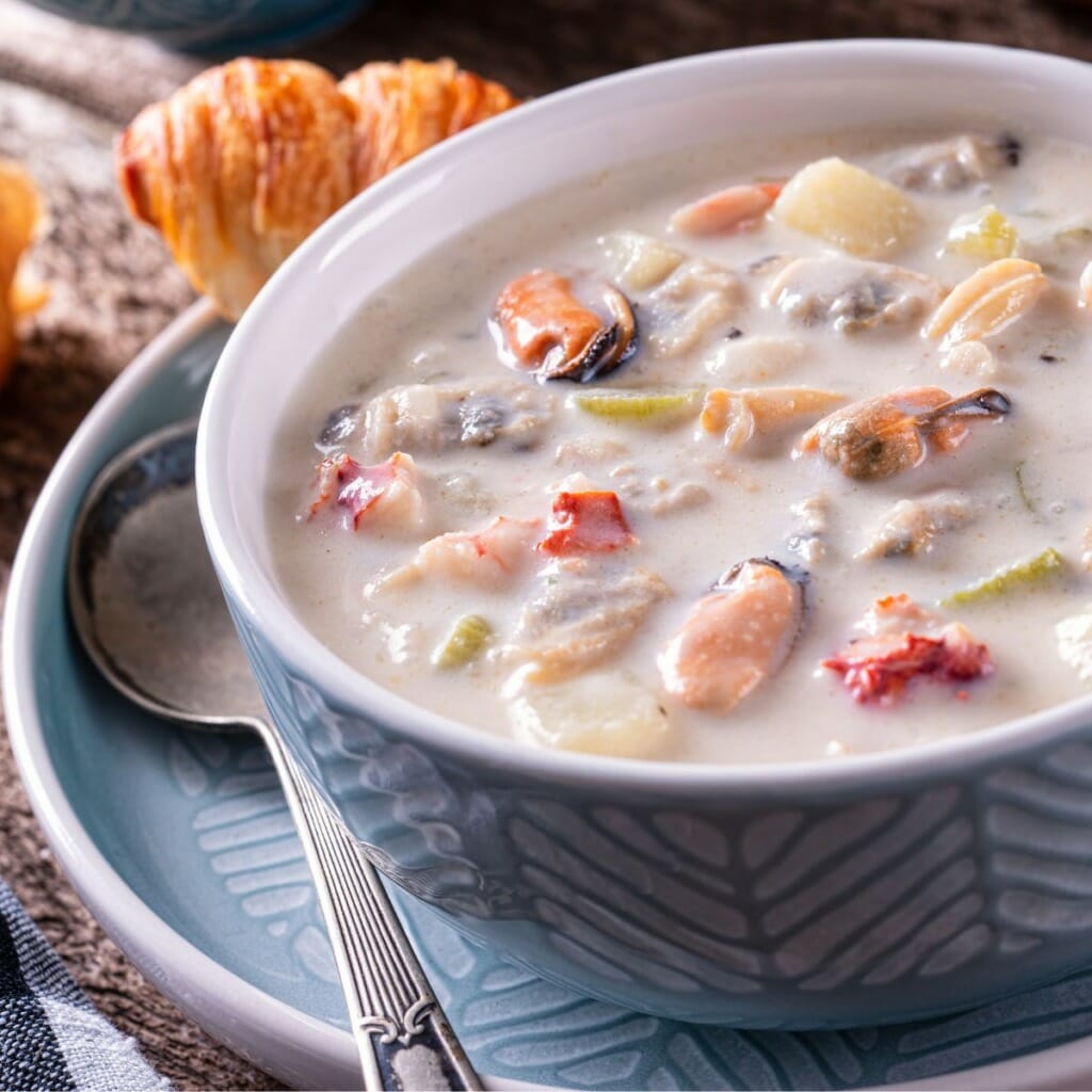 a bowl filled with seafood chowder with a metal spoon and a croissant
