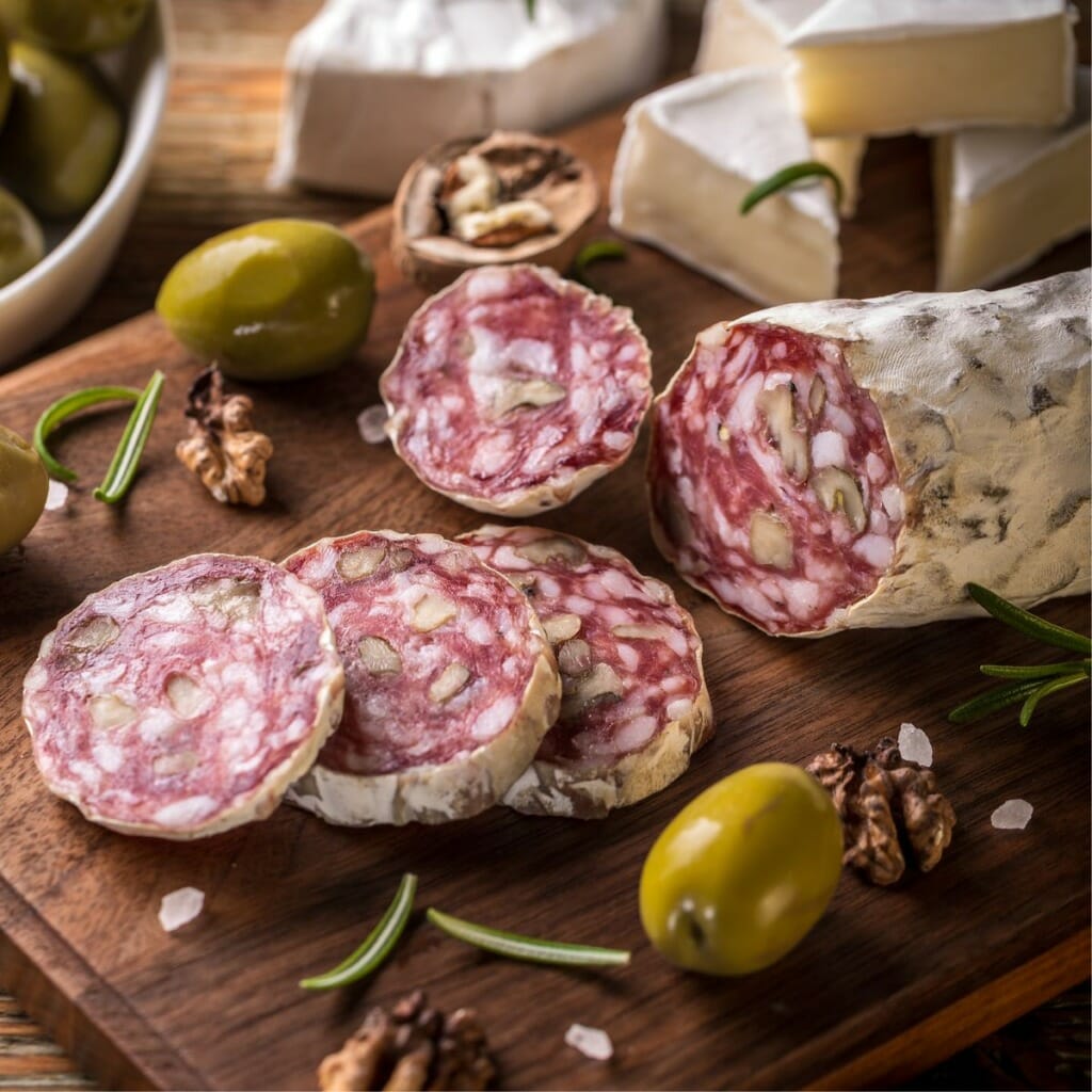 four slices of duck salami atop a wooden board, next to olives and rosemary
