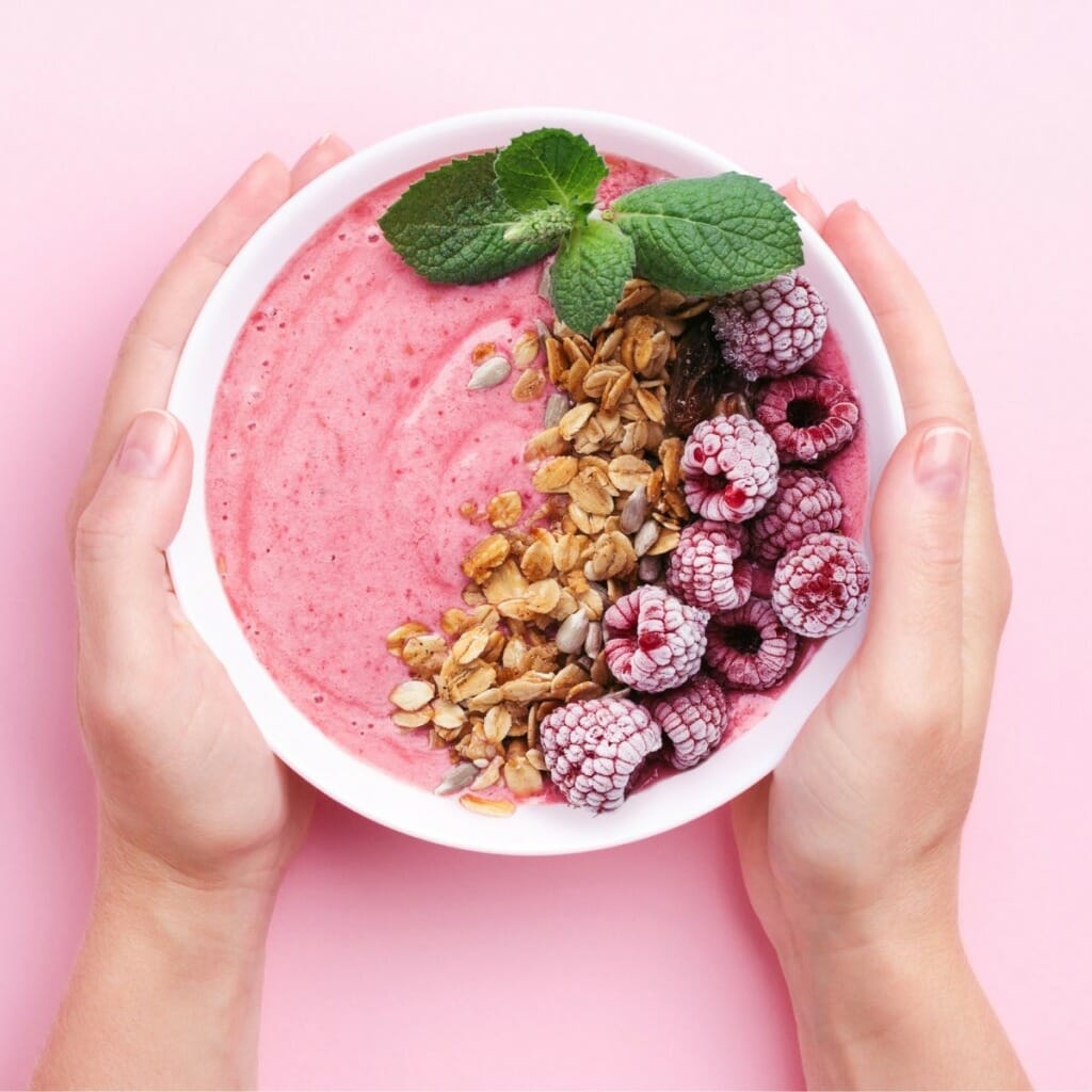 top view of hands holding a bowl of a pink smoothie topped with granola, raspberries, and a sprig of mint