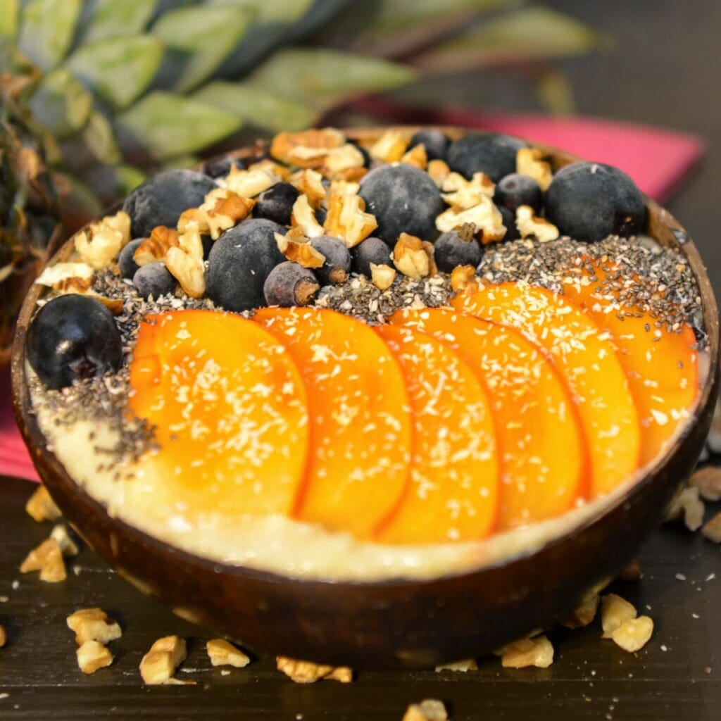 side view of a wooden bowl filled with orange smoothie, blueberries, chopped walnuts, chia seeds, sliced peaches, and shredded coconut