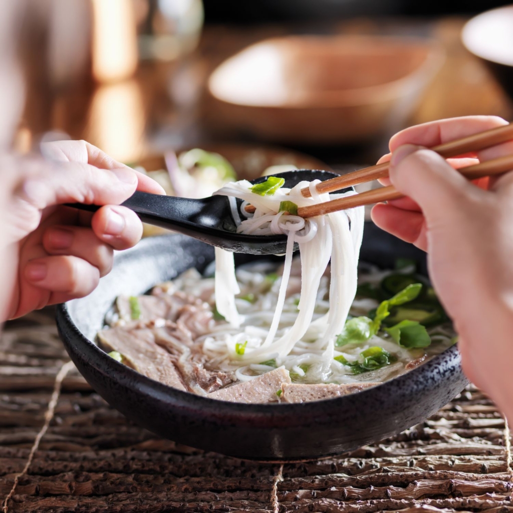 hands pulling pho noodles with chopsticks in the right hand and spoon in the left hand showing how to eat pho