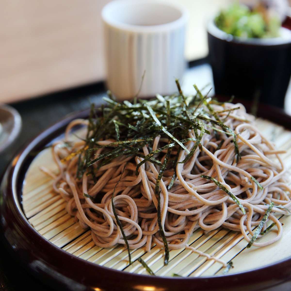 Soba noodles topped with greens in a plate in front of a cups on a table
