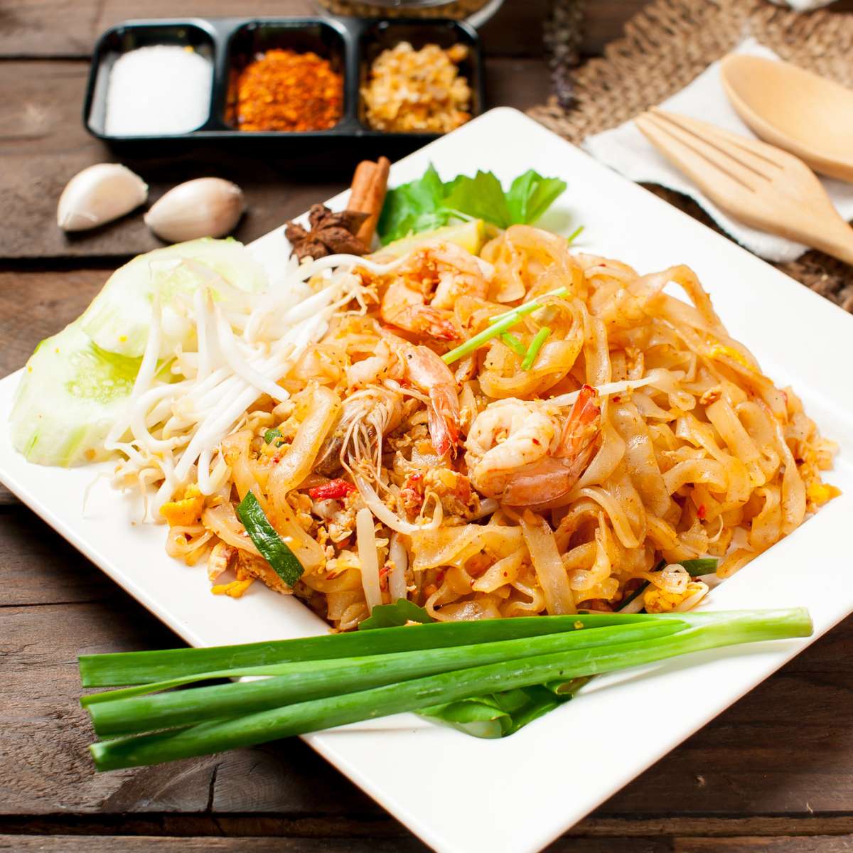 Egg noodles with prawn and vegetables in plate with spices, garlic clove, spoon and fork on a table 
