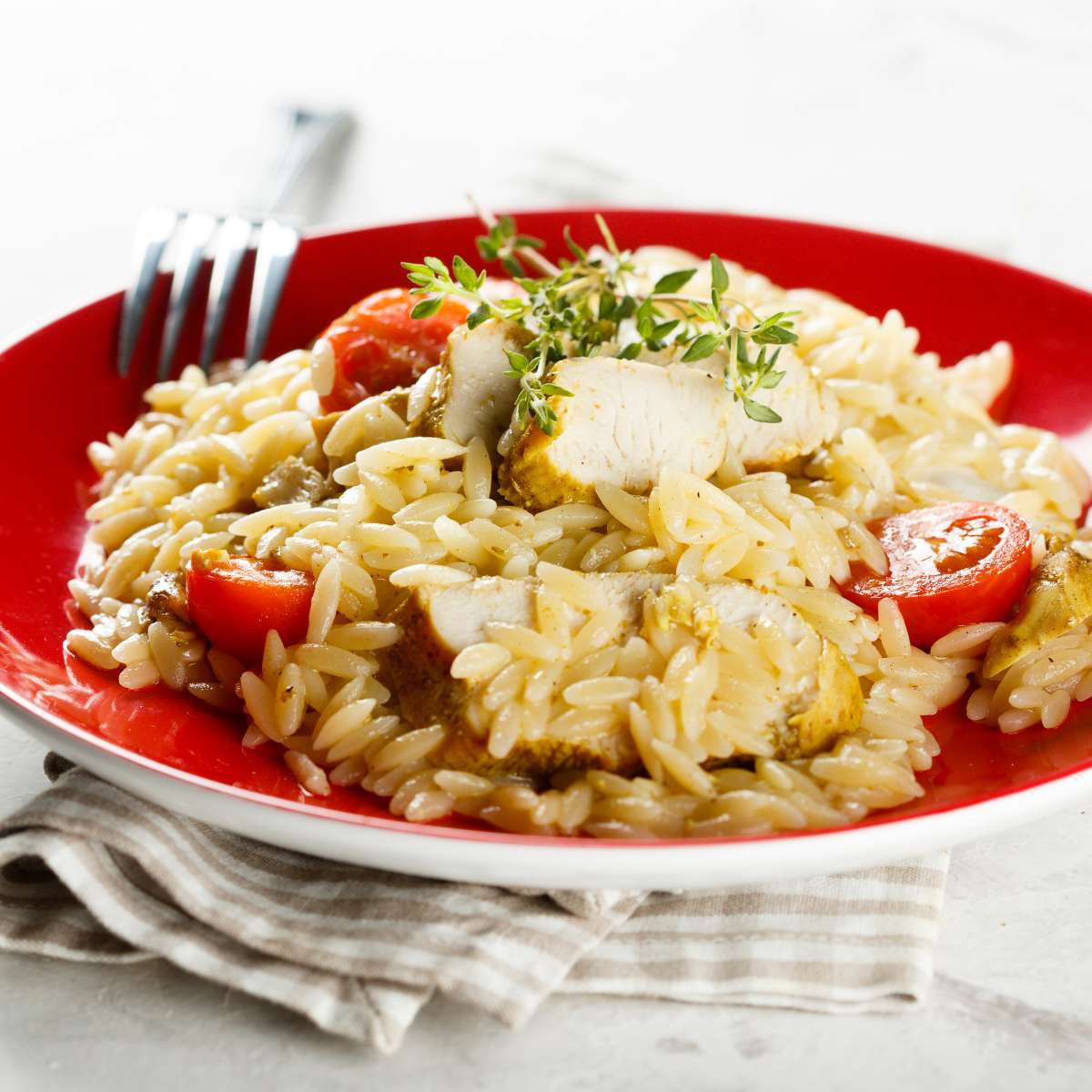 Orzo pasta with chicken, tomatoes and greens with a fork in a plate on a table
