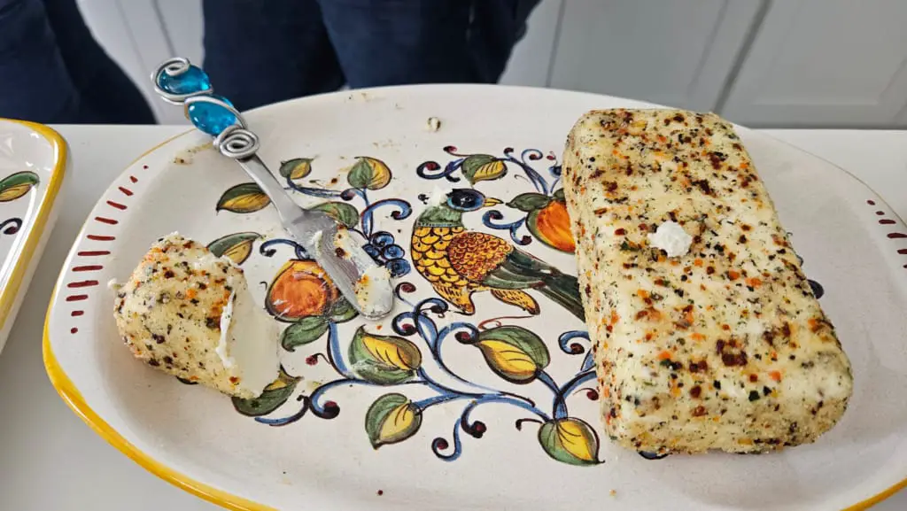 A loaf of goat cheese with herbs on a decorative plate, with a cheese knife.