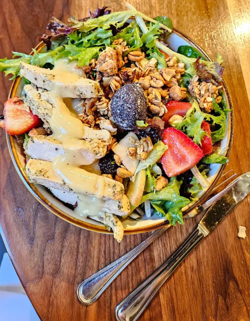 Strawberry arugula salad topped with granola crunch and burrata cheese covered in sesame seeds, and paired with grilled chicken on a plate alongside fork and knife on a table.