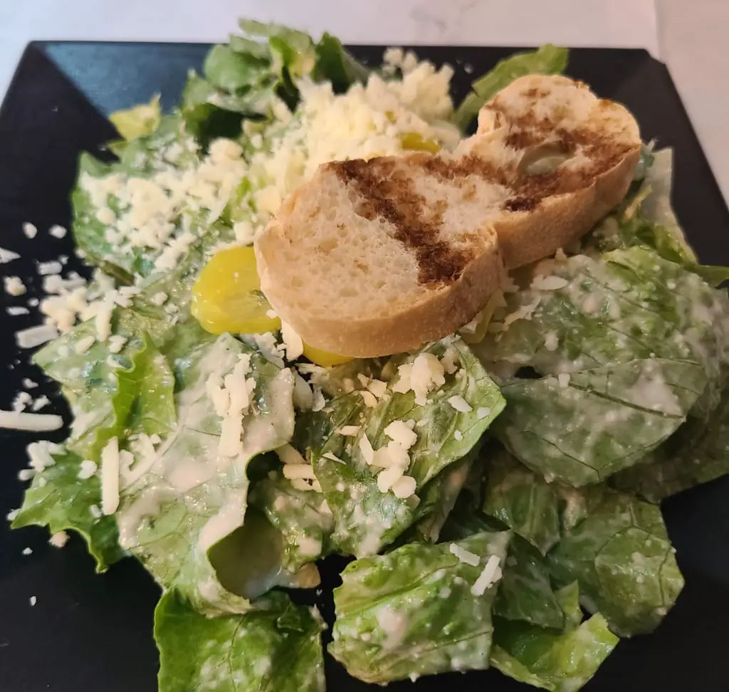 Caesar salad with lettuce and shaved cheese, served with a slice of toasted bread.