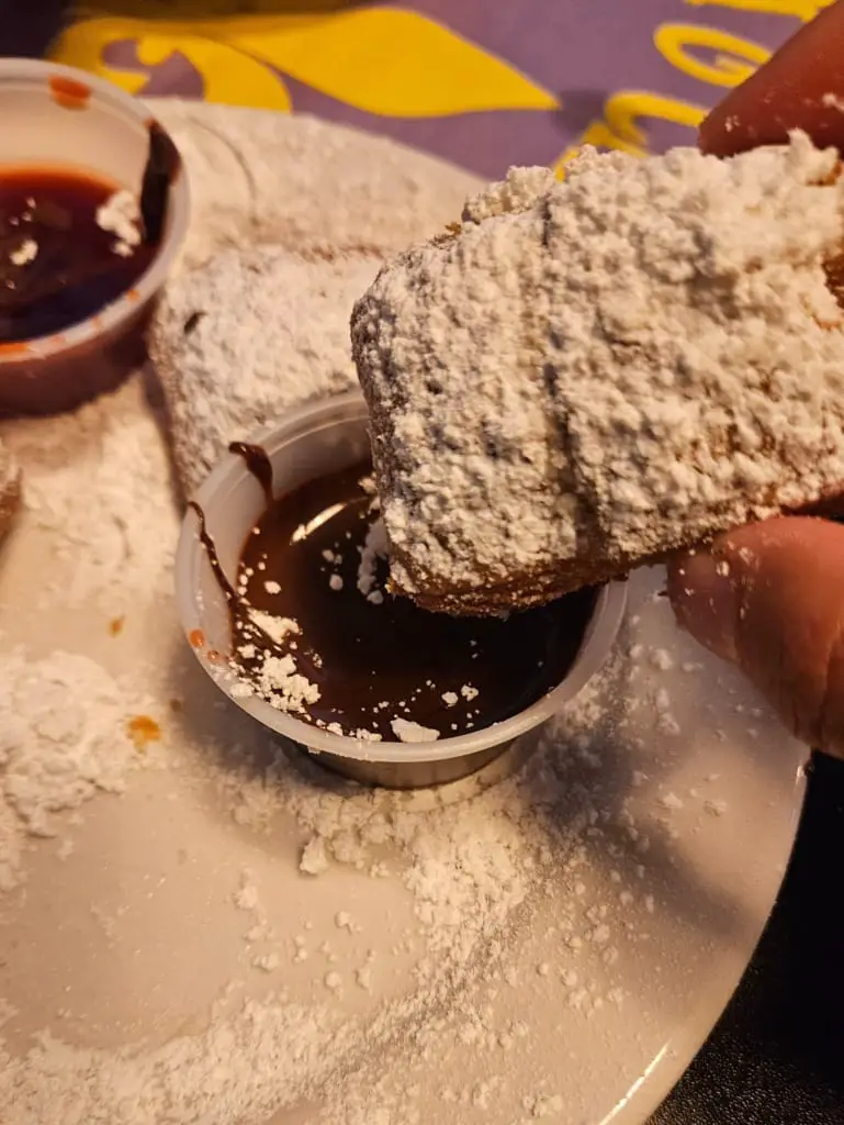 Hand dipping beignet covered in powdered sugar in chocolate.