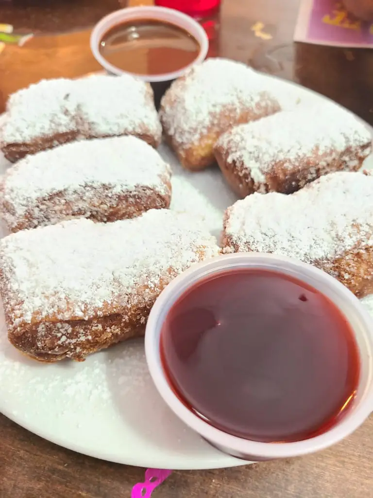 Beignets covered in powdered sugar on a plate with strawberry and chocolate dips.